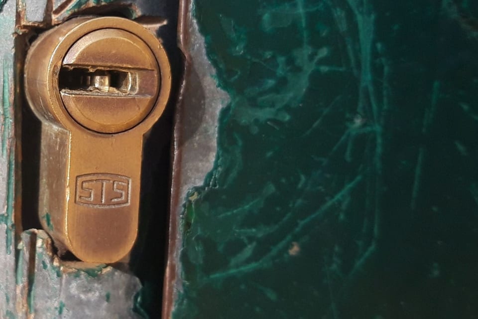 Close-up photograph of a brass euro-profile cylinder lock fitted through a chipped dark green painted steel door, keyway facing the camera.